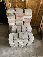 Stacked piles of used concrete pavers including rectangular unistone and flagstone types in gray, red, and beige colors.