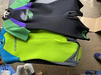 Top view showing folded Sea Doo women's wetsuit with black, green, and purple colors stacked on top of the unfolded neon green and blue O'Neill men's wetsuit.