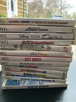 Stack of 13 Wii game cases showing Mario & Sonic at the Olympic Winter Games, Need for Speed Undercover, Disney Pixar Cars, Mario Super Sluggers, Wii Fit, Wii Play, Just Dance, Mario Kart Wii, NHL Slapshot, Big Beach Sports, Mario Power Tennis, Super Mario Galaxy 2, Winter Sports.
