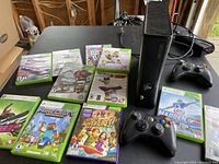 Xbox 360 console in upright position with 2 wireless controllers and 11 game cases laid out on table.