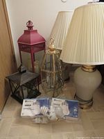 View of red lantern, smaller brownish-black lantern, large crystal lamp, two cream-colored lamps with beige shades, and package of emergency light bulbs on the floor.