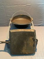Photo showing top view of the square cast iron skillet and the Dutch oven pot with lid in the background. Skillet shows signs of discoloration and use.