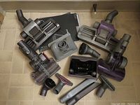 Photo showing eleven Dyson vacuum attachments of various types including motorized heads, brushes, and nozzles in gray, black, and purple plastic.