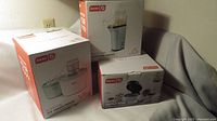 Three boxed Dash appliances including ice cream maker, waffle bowl maker, and popcorn maker on a gray surface against a beige wall.