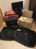 Overview photo showing all items: two Samsonite briefcases (beige-gold and black), black cosmetic bag, bright orange roadside kit pouch, black neck pillow, two zipped pouches, and three luggage straps.