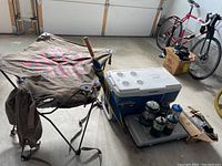 Photo showing cooler, foldable camping table with umbrella, frying pans, Coleman lantern and stove together in garage setting.