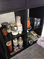View of multiple scented candles in jars, tins, and containers on lower shelves of black shelving unit.