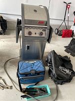 Coleman gas BBQ grill with cooler bag, BBQ cover, and small container of BBQ accessories arranged in a garage setting.