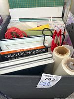 Photo showing a variety of craft supplies including adult coloring books with animal designs, red and black cords, rolls of tape, paper doilies, and craft bags.