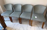 Four upholstered dining chairs in light green fabric with wooden legs, arranged side by side on a tiled floor.