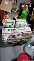 Overview of FoodSaver vacuum sealing system box with multiple boxes of vacuum sealing bags visible, background contains unrelated games and toys.