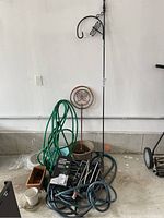 Setup of garden hoses coiled on floor near plant pots and shepherd's hook against garage wall for a broad overview.