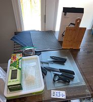 Wide view of kitchenware lot showing cutting boards, knives with covers, peeler, placemats, vented containers, and cookbook stand.