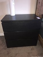 Front view of the black three-drawer nightstand showing the smooth finish and drawer fronts with recessed handles.