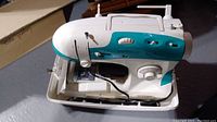 Compact Singer sewing machine with white and teal body, with control knobs and electrical cord.