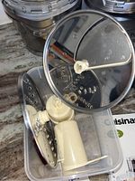 Close-up photo of food processor parts, including stainless steel slicing and shredding discs and plastic components inside a clear plastic container.