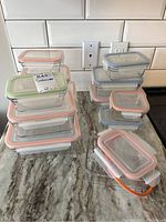 Photo of a collection of clear rectangular glass food storage containers stacked on a granite countertop, each with colored silicone-sealed lids in orange, green, and blue. One extra round silicone seal is visible.