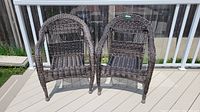 Two brown cane patio chairs positioned side by side outdoors on a deck, showing front and slightly angled view, highlighting their woven design and minor wear.