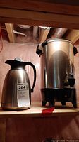 Photo of a stainless steel coffee carafe with black handle and coffee urn in a basement setting.