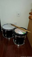 Pair of black snare drums side by side on floor with pair of wooden sticks resting on top.