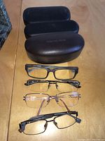 Four pairs of eyeglasses displayed horizontally with three black glasses cases behind them, two cases labeled Michael Kors.