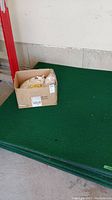 Photo of five stacked green golf driving mats with an open cardboard box on top containing white rubber tee holders.