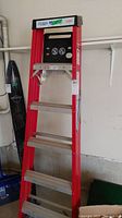 6 ft metal step ladder with red rails and aluminum steps photographed leaning against a garage wall, showing black plastic top tool tray with slots and holes