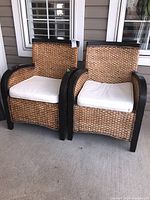 Two matching outdoor wicker patio chairs with removable white cushions sitting side by side on a porch with window shutters behind.