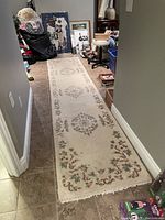 Photo of long light beige runner rug placed in narrow hallway with tile floor. Shows floral pattern with central medallions and floral borders. Fringe visible at near edge.