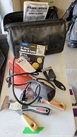 Photo showing the Black and Decker orbital sander, carrying bag, sanding discs, instruction manual, and scrapers arranged on a white surface.