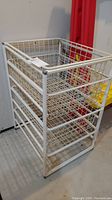 White metal wire storage shelf with three basket-style tiers, set against a gray wall and floor with a red pillar to the side. Basket shelves are removable and have a grid wire pattern.