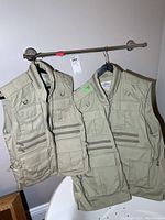 Two beige Tilley 15-pocket outdoor utility vests hanging on a rod, showing front view with multiple zippered and flap pockets, side adjustment straps, and hanging tags.