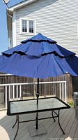 Front view of the large blue vented patio umbrella fully opened on a glass patio table, showing the tiered canopy design and black metal pole.