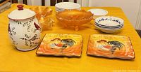 Overview of all glass and ceramic dishes on a yellow tablecloth including rooster plates, carnival glass dishes, white plates, and covered jar