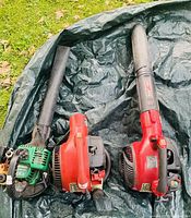 Three gas-powered leaf blowers placed in a tarp, two red Troy-Bilt and one green with parts visible.