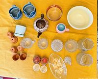 Top down image showing all items laid out on a yellow cloth including ceramic pots, pitchers, teapot, small glass candle holders, and glass vases