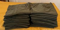 Two photos showing a stack of 23 folded dark green US Army tropical pants size 28XL, with side cargo pockets and standard pockets.