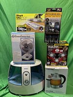 Photo shows all items in the lot grouped together including the Vicks humidifier, boxed Windmere fan, boxed Shark VacMop pads, boxed Tri-Burst LED light, boxed Rattle Snake Coil cable, and boxed Chefman kettle.