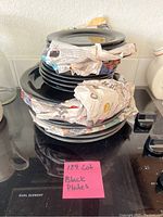 Stacked black plates of various sizes with some wrapped in newspaper on black countertop.