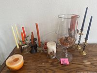 Close-up of cedar wood surface with some candles and two large glass hurricane candle holders containing decorative filler and pine cones, assorted colorful tapered candles in holders.
