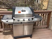 Photo showing a full view of a Charbroil stainless steel gas grill with three control knobs and side burner knob, placed outdoors on a deck. Grill has stainless steel doors and side shelves, appears heavily used and needs cleaning.