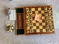 Photo showing complete chess set with brass chess pieces on a solid wood chessboard with green felt storage drawers and extra game pieces including checkers and dominoes in bags