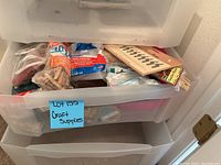 View of open plastic storage drawer showing assorted craft supplies including glue sticks, wooden clothespins, and other items