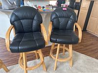 Pair of black leather swivel bar stools with light wooden arms and base shown from front on wood floor near kitchen counter.