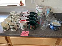 Photo showing various Christmas-themed mugs with tree and wreath patterns, clear glasses with holiday designs, and some Christmas candle holders.