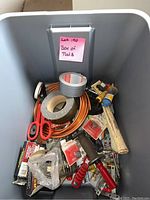 Photo of storage bin showing assorted hand tools including scissors, duct tape, clamps, small hardware items, and copper piping.
