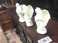 Four white ceramic musical angel candle holders arranged on a wooden cabinet.