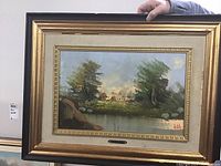 Framed landscape painting showing trees, water, and buildings with a gold-tone frame.
