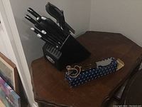 Full view of black knife block with multiple kitchen knives with black handles, a custom knife in blue box, and a manual sharpener on wooden table.