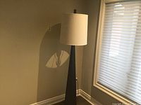 Full view of the floor lamp showing black tapered metal base and large round off-white lampshade against a beige wall and window.